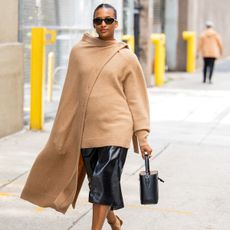 A woman wearing a tan scarf sweater, a leather skirt, and a black bucket bag.