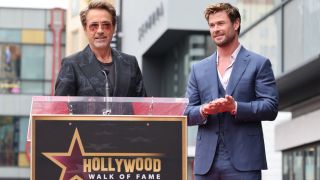 Robert Downey Jr. speaking at a podium while Chris Hemsworth happily watches on, during his Hollywood Walk of Fame ceremony.