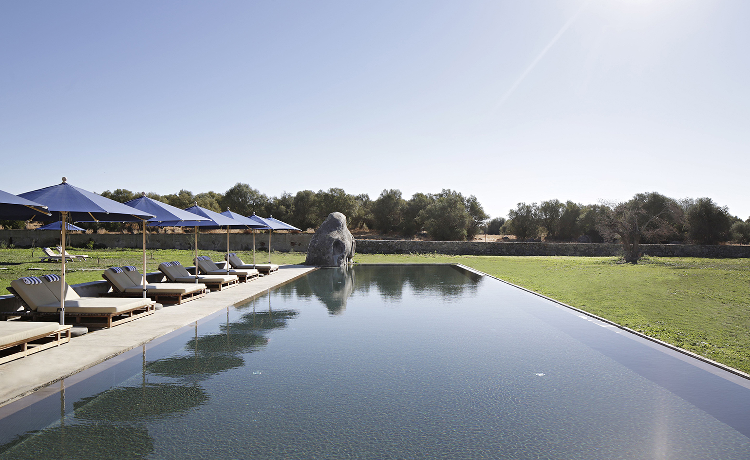 A pool area in the São Lourenço do Barrocal hotel. At the center is the pool with a rock-like fountain in the left corner. To the left, we have beach beds in beige and navy sunshades.