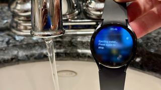 Samsung Galaxy Watch 6 next to a bathroom water faucet, showing the Water Lock info screen: "Ejecting water. Press again to stop."