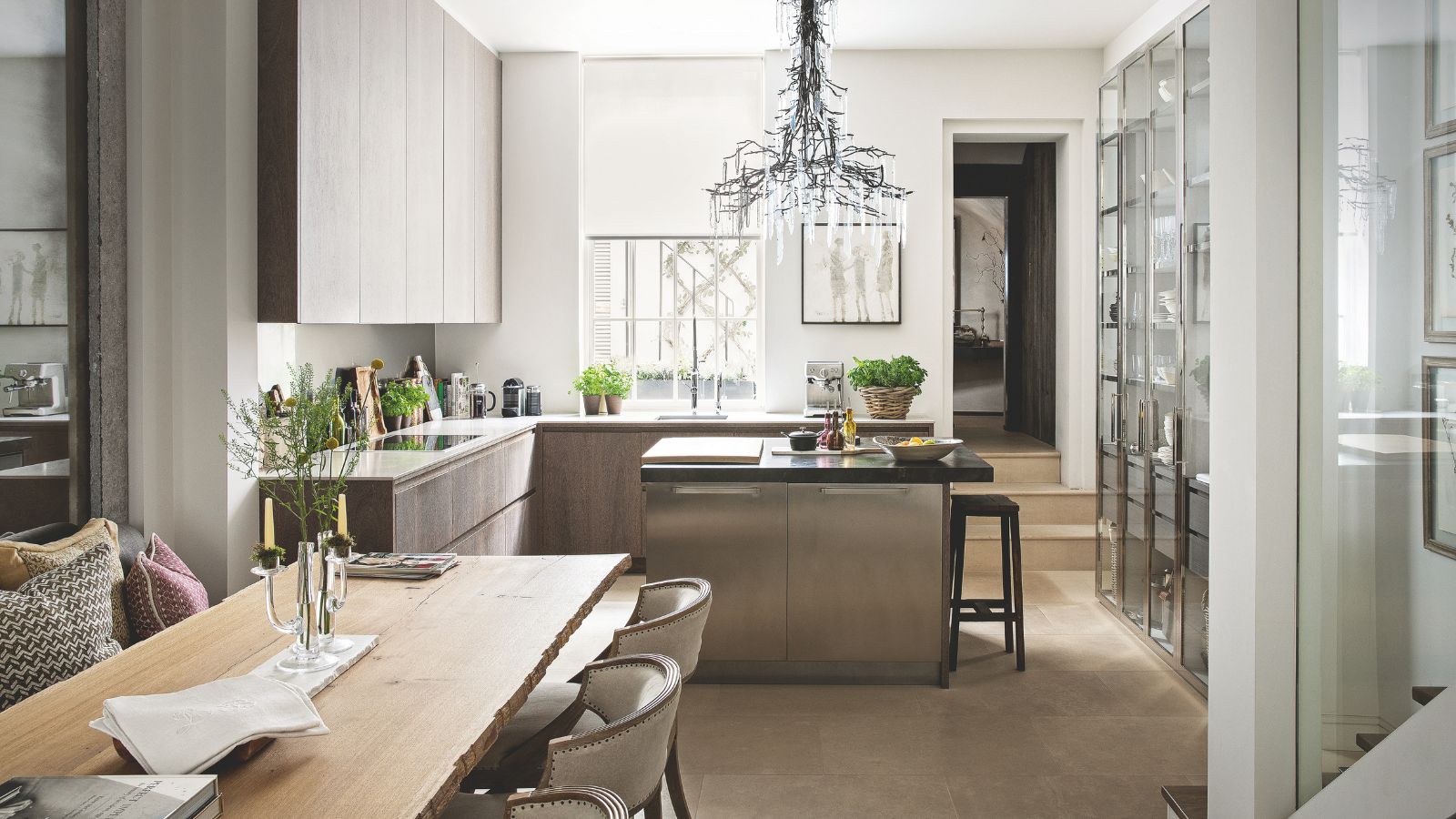 A neutral kitchen with tiled flooring and white walls. In the center is an island with stool seating, and around it are wooden cabinets and white countertops. To the right are glass-fronted cabinets, and in the foreground is a wooden dining table with white chairs around it.