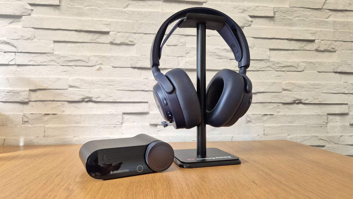 The SteelSeries Arctis Nova Pro Omni headset with its GameHub sitting on a headset stand in front of a white brick background and on a wooden surface.