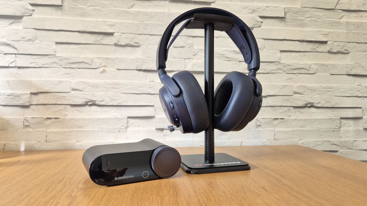 The SteelSeries Arctis Nova Pro Omni headset with its GameHub sitting on a headset stand in front of a white brick background and on a wooden surface.