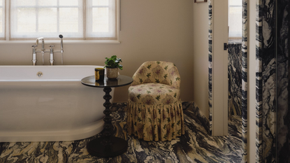 a marble bathroom with bobbin side table next to a freestanding bath, and yellow armchair with patterned skirt
