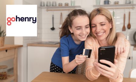 Daughter holding her Gohenry debit card leaning on her mother who is smiling at something on her phone