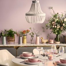 Pastel pink dining set on round table with coloured glasses and burning candle next to ornaments on on sideboard
