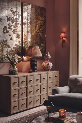 A red living room with grey armchair and antique sideboard. There is a large table lamp switched on, along with a wall lamp.