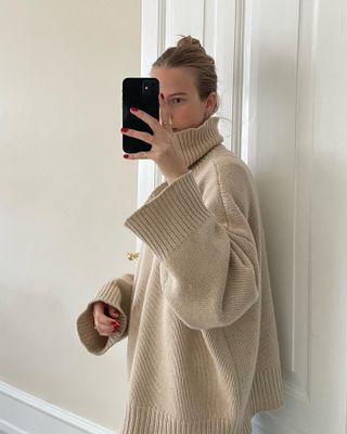 Scandinavian style influencer and creative Clara Dyrhauge poses for a mirror selfie wearing a winter outfit with a twisted updo hairstyle, red nails, and a beige oversize turtleneck sweater