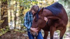 Woman and her bay horse in a forest. The horse is bending his head towards her and she has her arms around him.