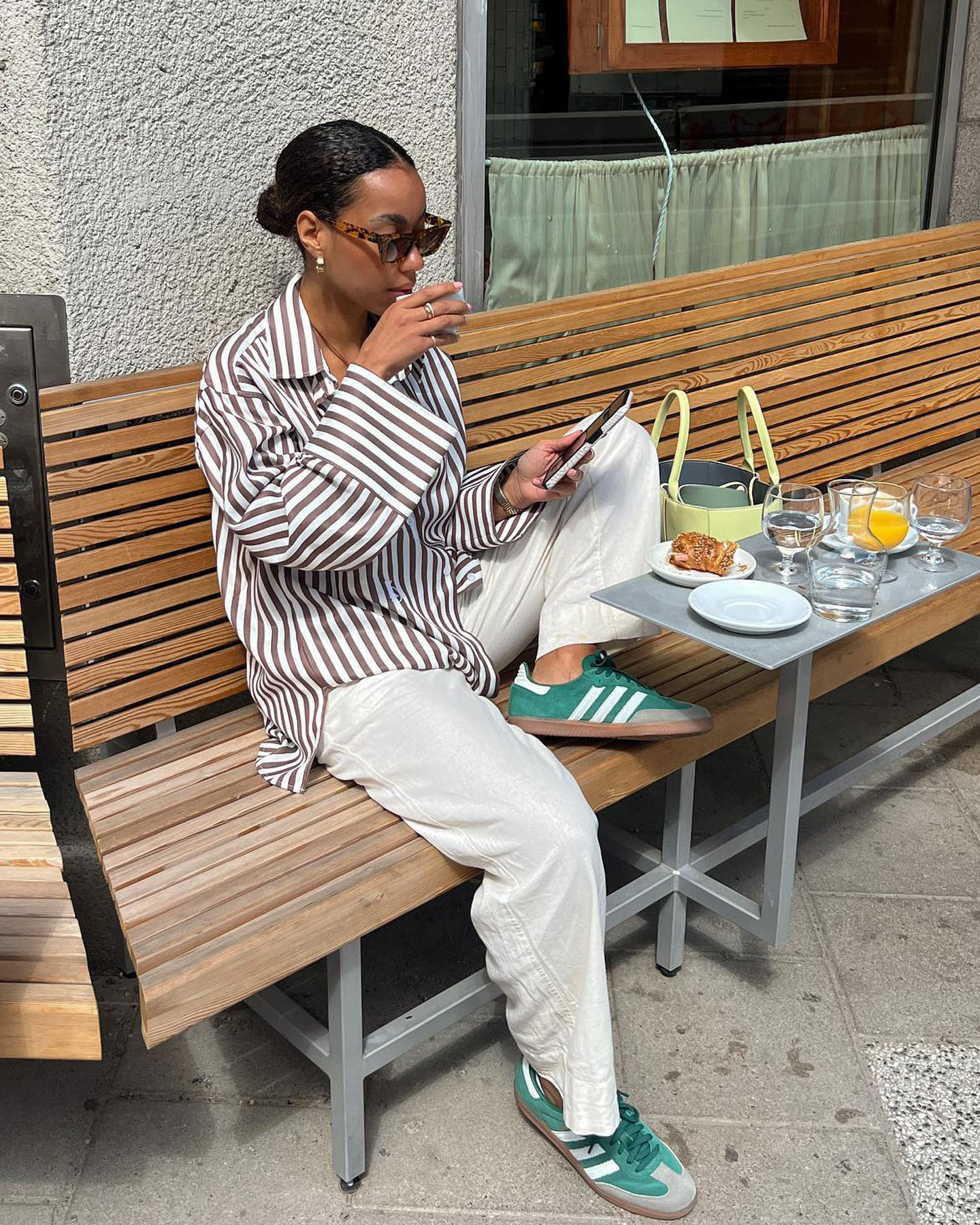 stylish woman sits at an outdoor cafe wearing tort sunglasses, a brown and white striped button-down shirt, linen pants, and green Adidas Samba sneakers