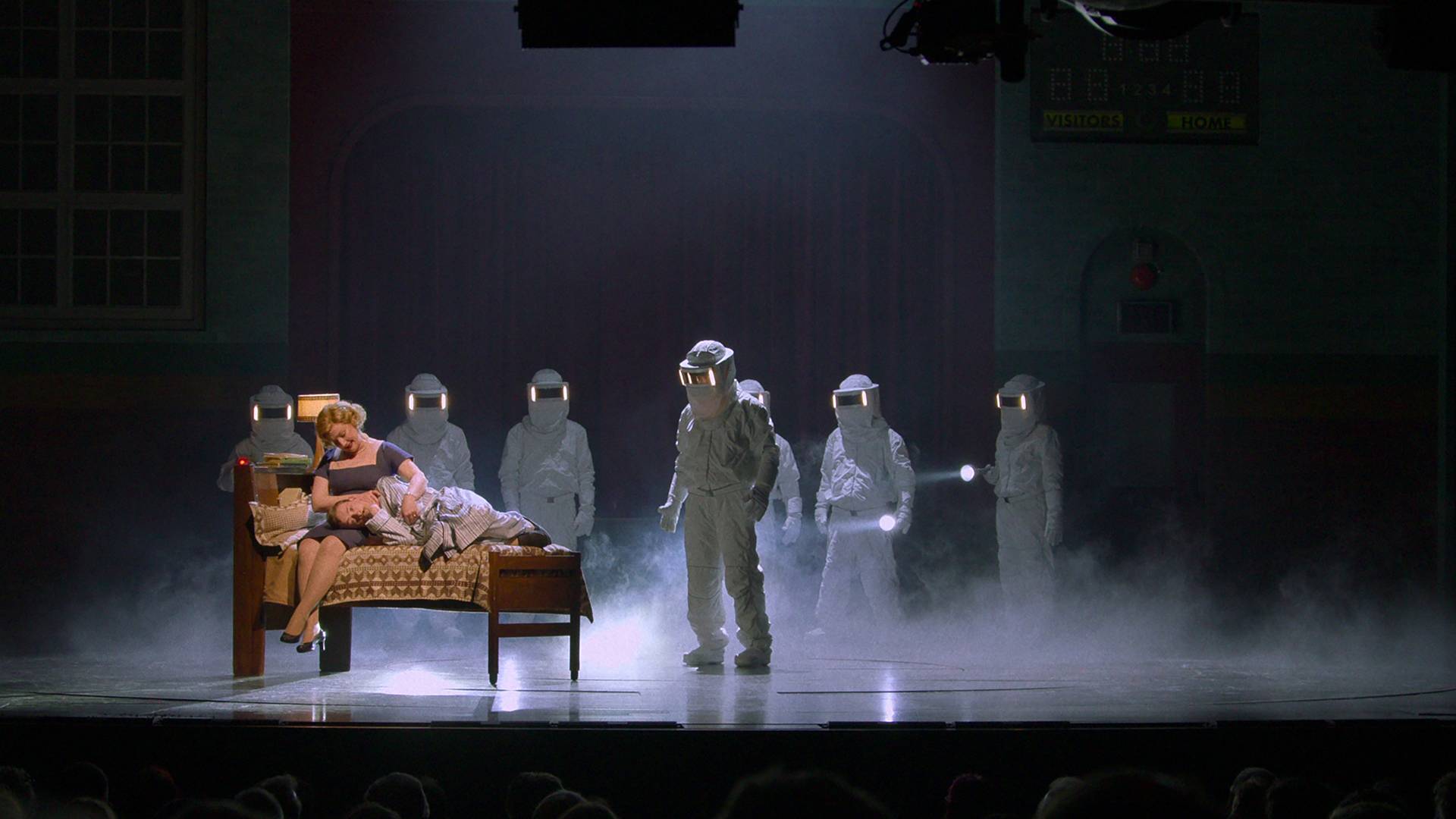A still image from the play Stranger Things: The First Shadow, featuring people in hazmat suits and a woman holding a child on a bed