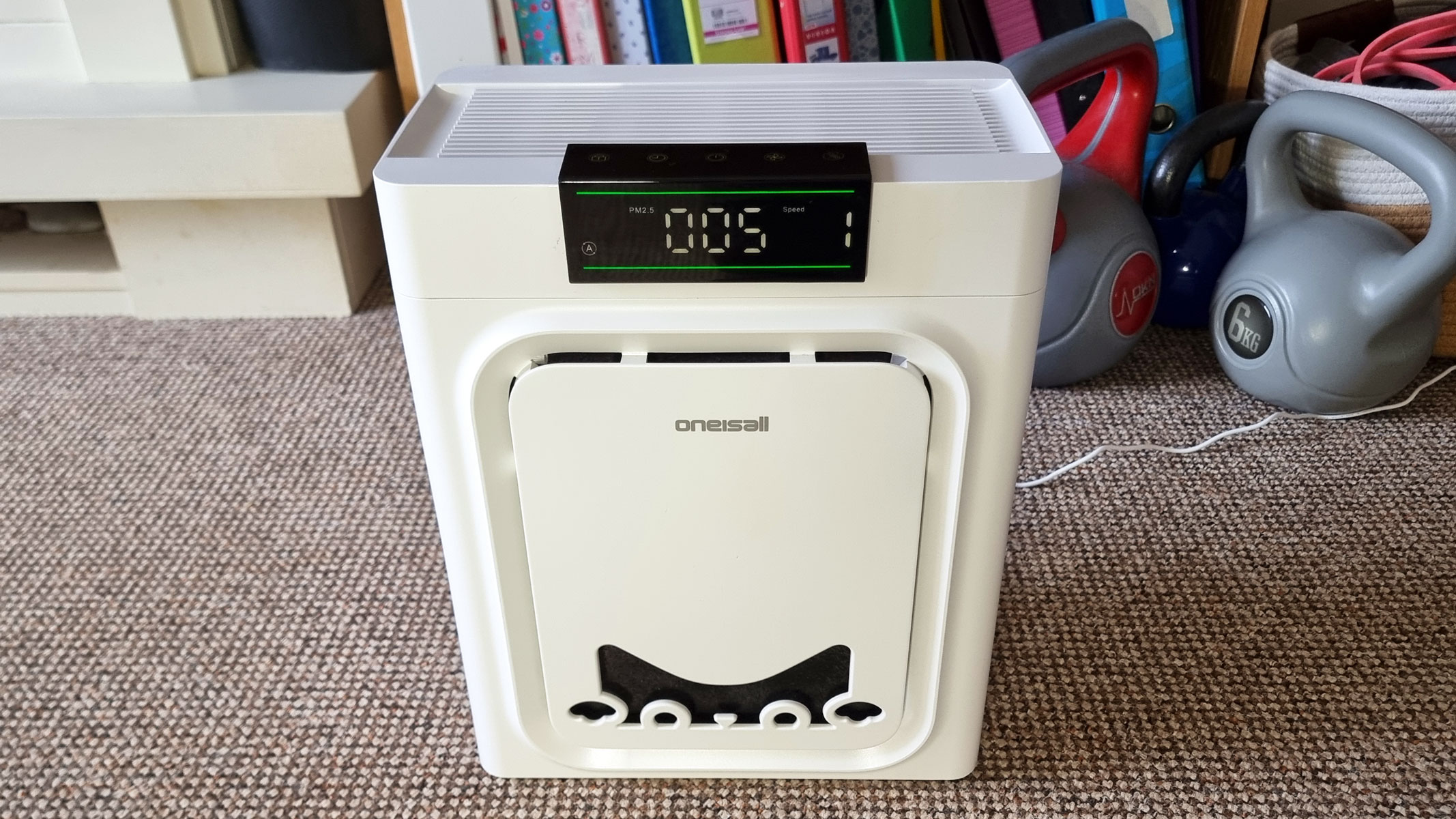 a picture of the Oneisall air purifier standing in front of a bookshelf and a pair of kettlebells
