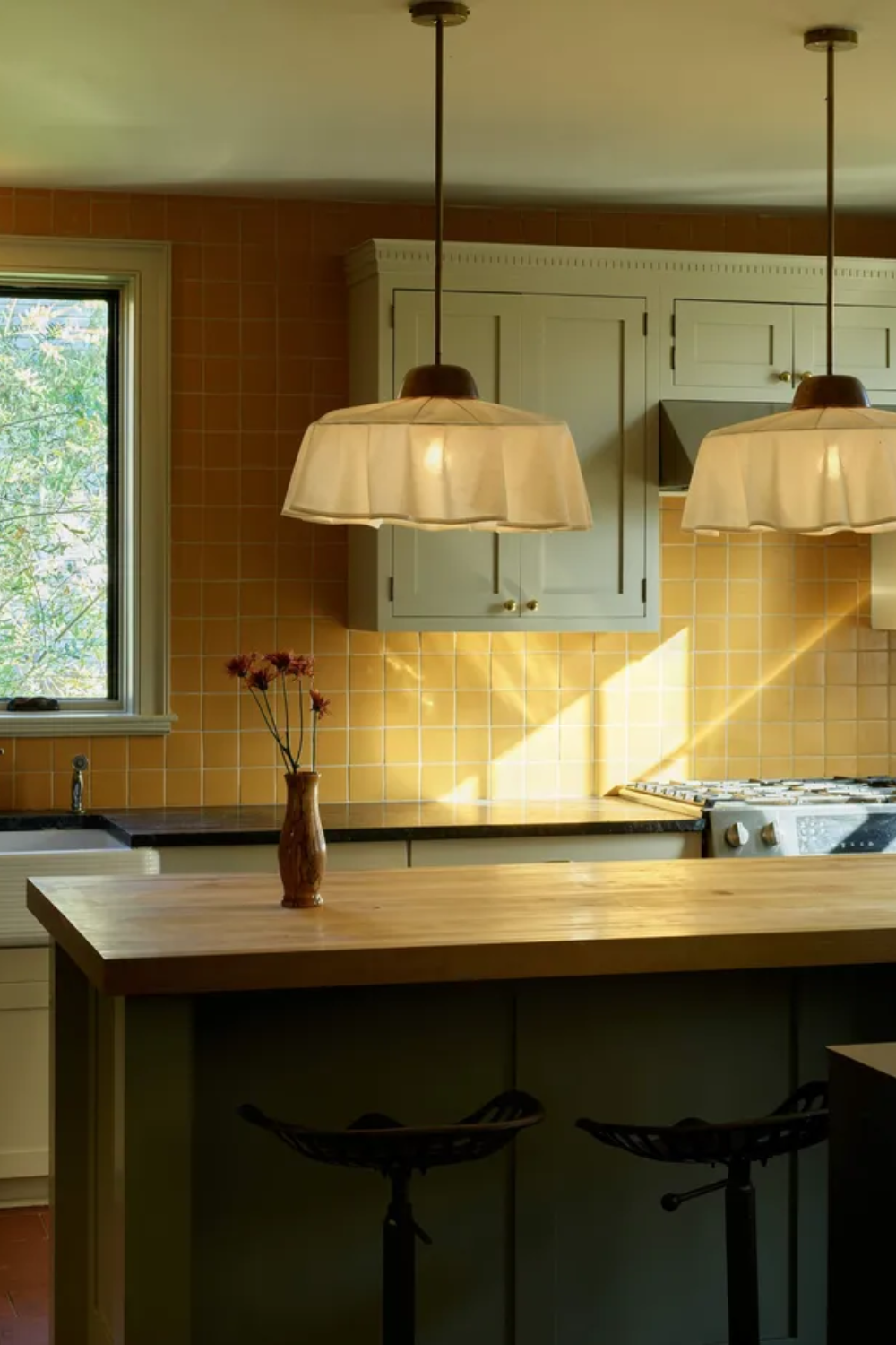 Kitchen with light wood butcher block counter, two white ruffled pendant lights, off-white upper cabinets, and a backsplash made of small, square orange/yellow tiles