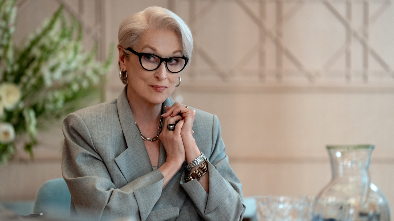 Miranda Priestly sits behind a desk with her hands clasped, commanding attention in a grey suit and glasses in the Devil Wears Prada 2.