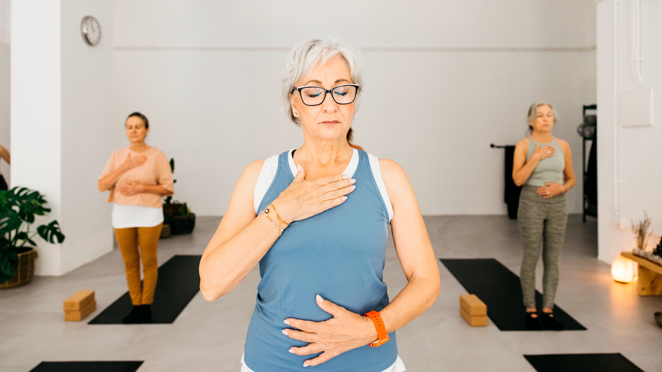 Woman standing in yoga class with one hand on her breastbone and one hand on her stomach. Her eyes are closed. Two women stand either side of her in the background, in the same position.