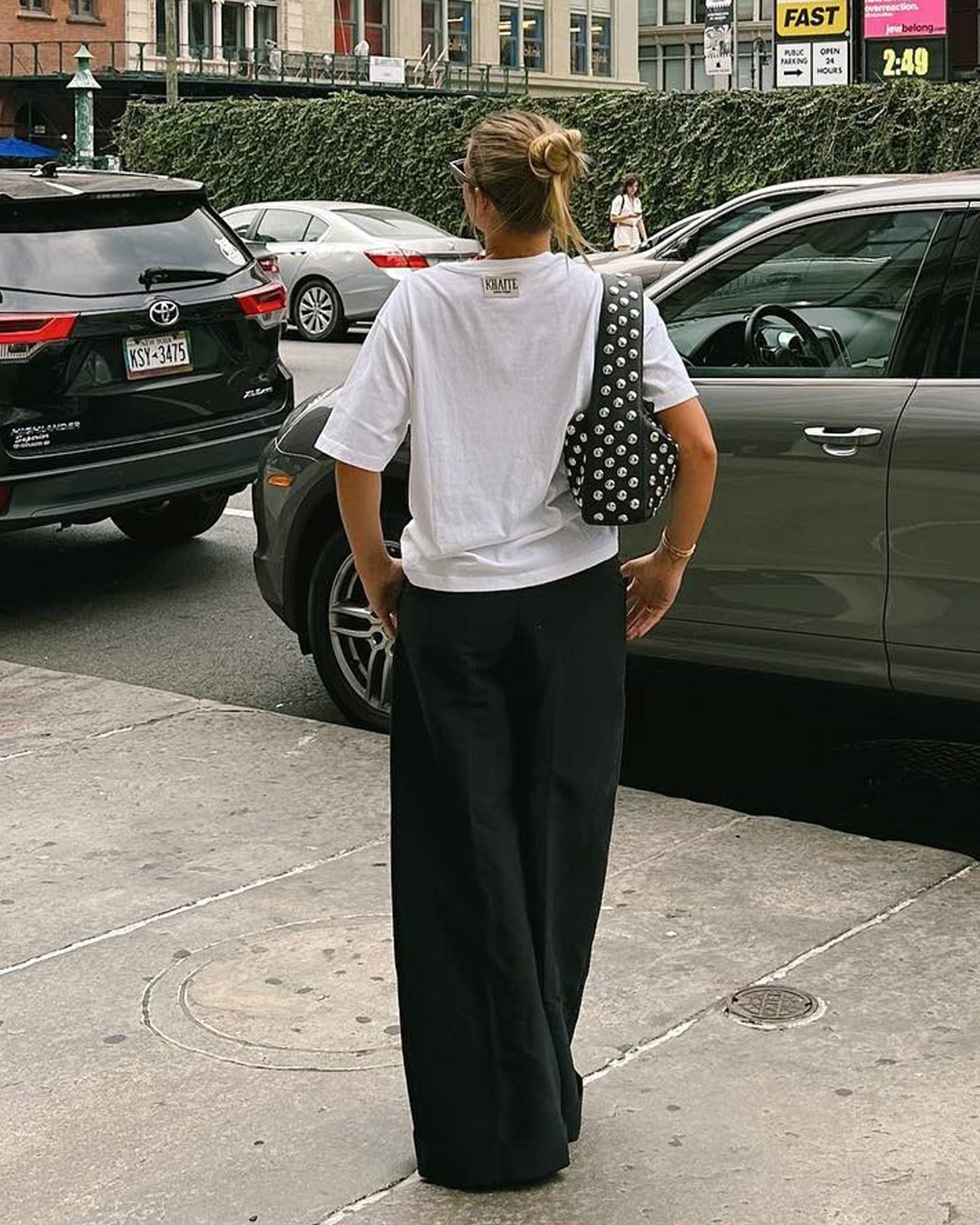 Sofia Richie Grainge stands on a sidewalk in NYC wearing a studded black shoulder bag, oversize white t-shirt, and wide-leg black pants.