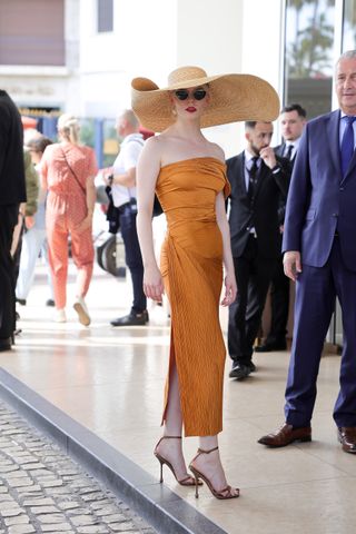 Anya Taylor-Joy in Cannes wearing a big hat and orange strapless dress and high heels.