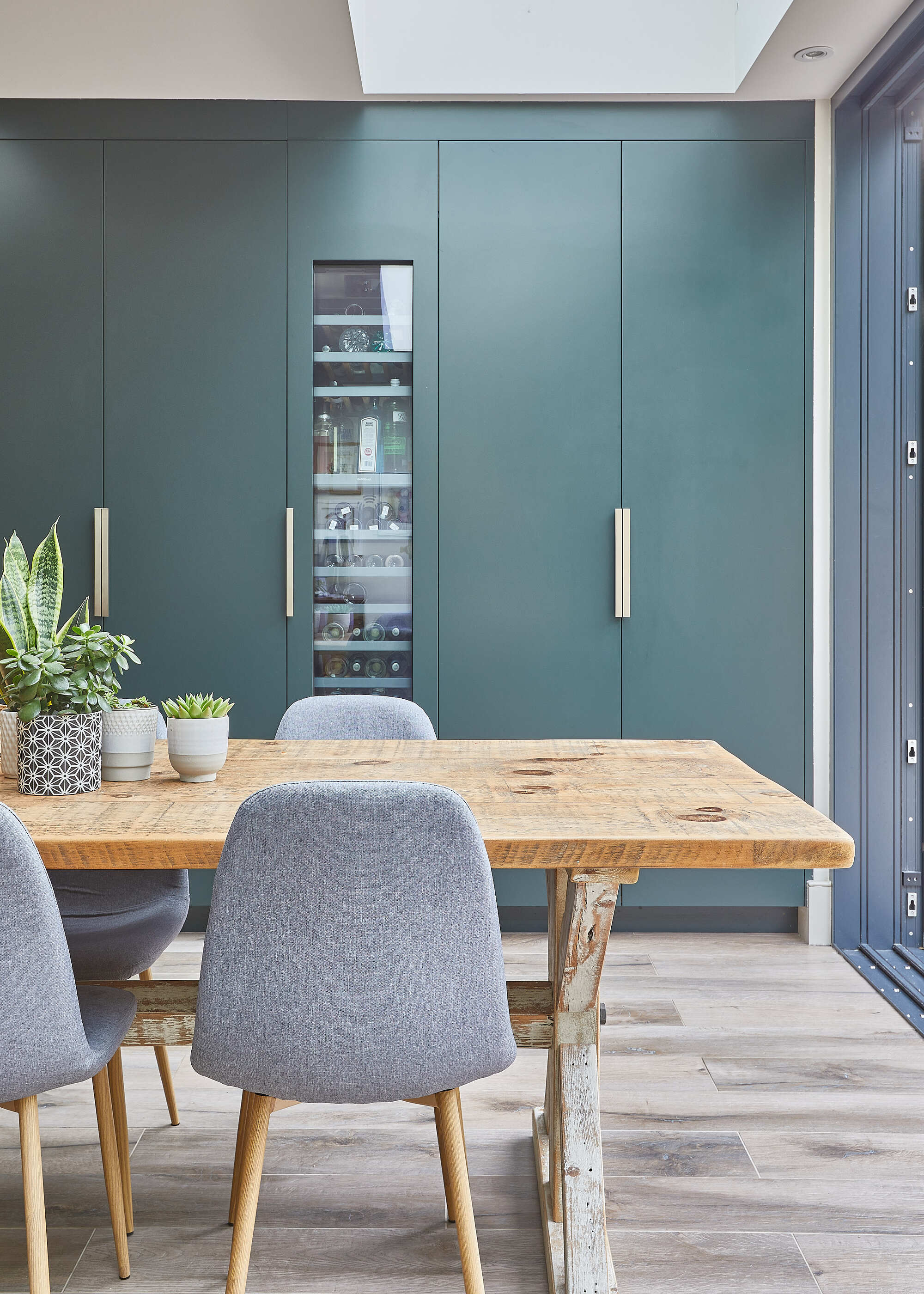 A kitchen with a large wooden dining table, wooden flooring, and floor-to-ceiling teal cabinetry with a wine fridge built in