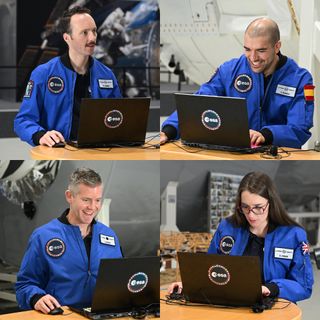 four people in blue flight suits laugh and smile as they play a computer game on four separate laptops