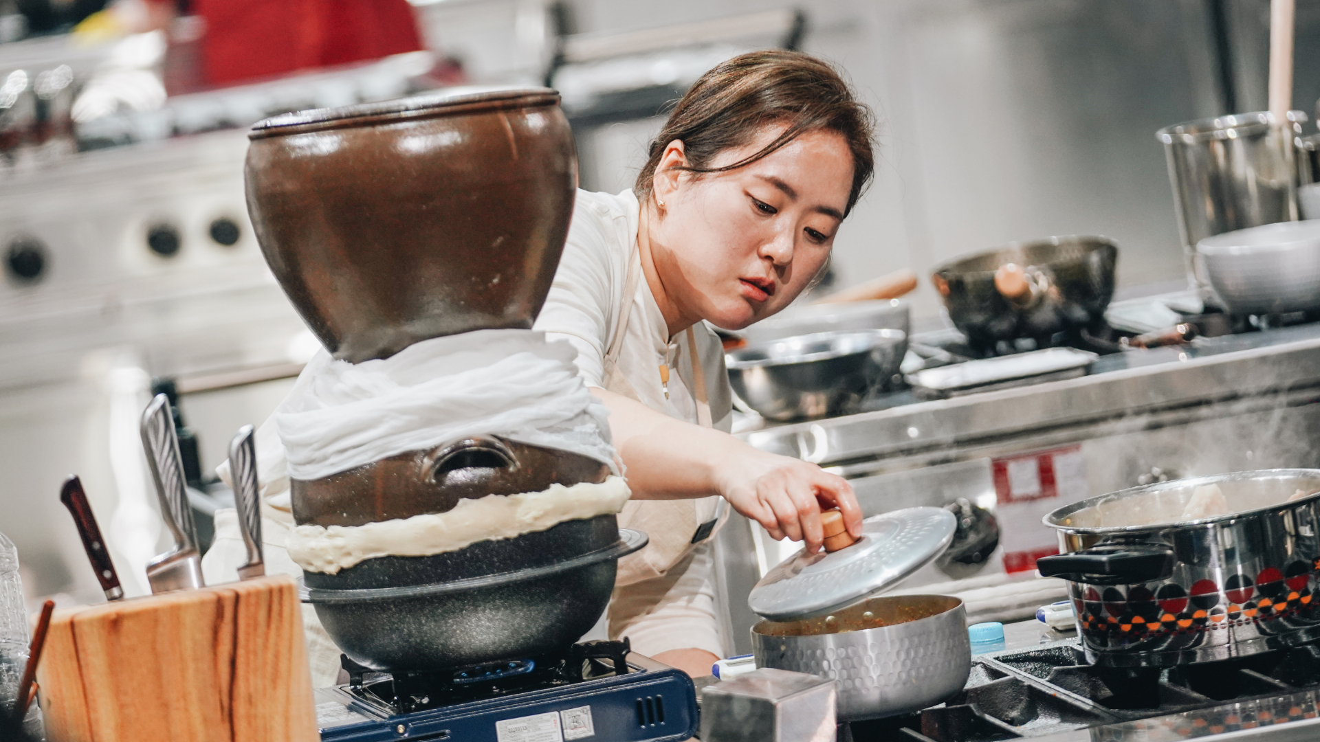 A female chef nicknamed Brewmaster Yun lifts a lid on a small pot while standing at a workstation with a traditional liquor distilling pot, in 'Culinary Class Wars' season 2.