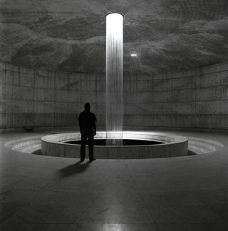 Interior of the Museum of Independence, Dhaka. The photo is black & white. The photo shows a man standing next to a column of water that's cascading from the ceiling. The room is circular.