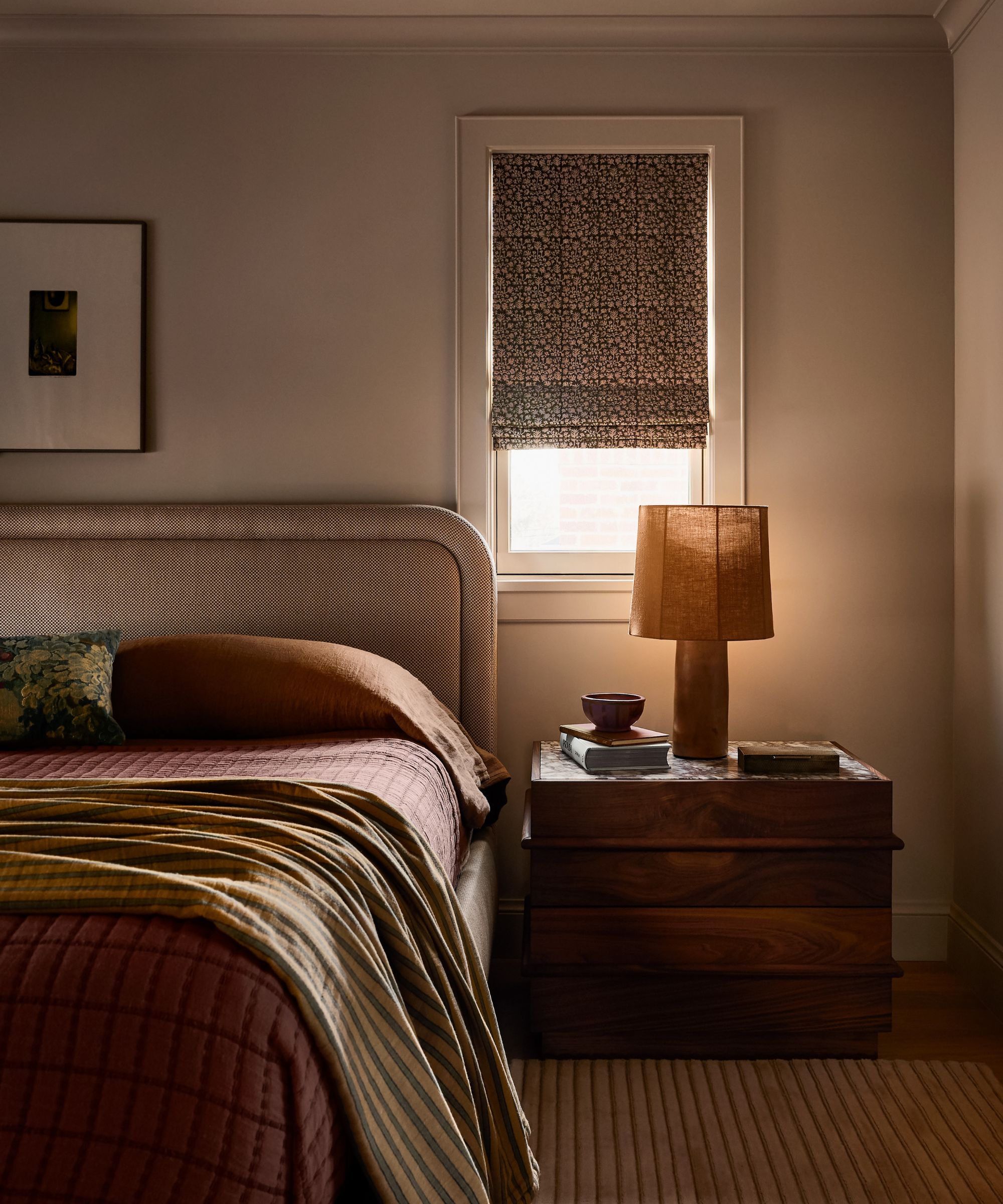 an earthy primary bedroom with small window, upholstered beige rounded headboard, wooden nightstand, striped rug and muted red bed linens