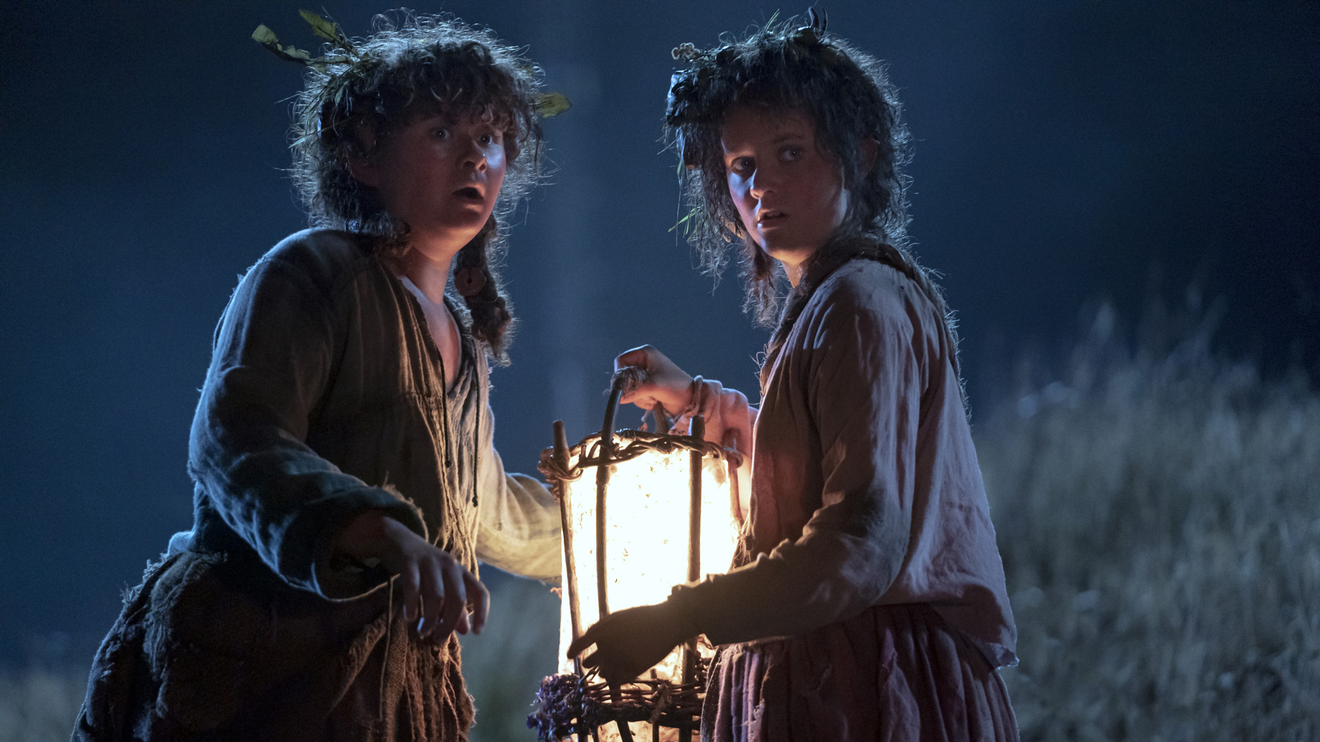 Nori Brandyfoot and Poppy Proudfellow look behind them with their faces lit by a lantern at night in The Rings of Power