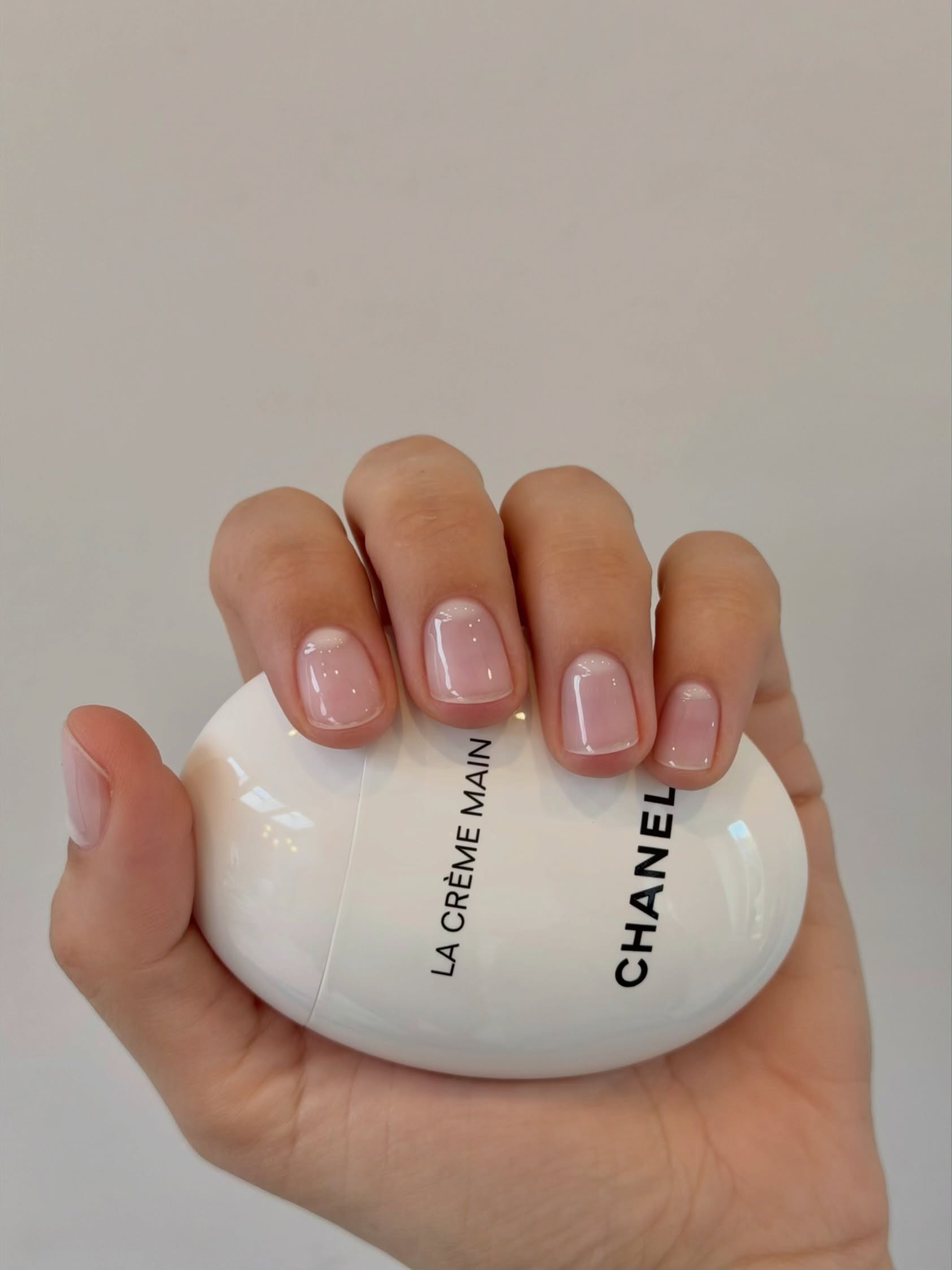 A woman's hand holding a Chanel hand cream with clear, glossy nails