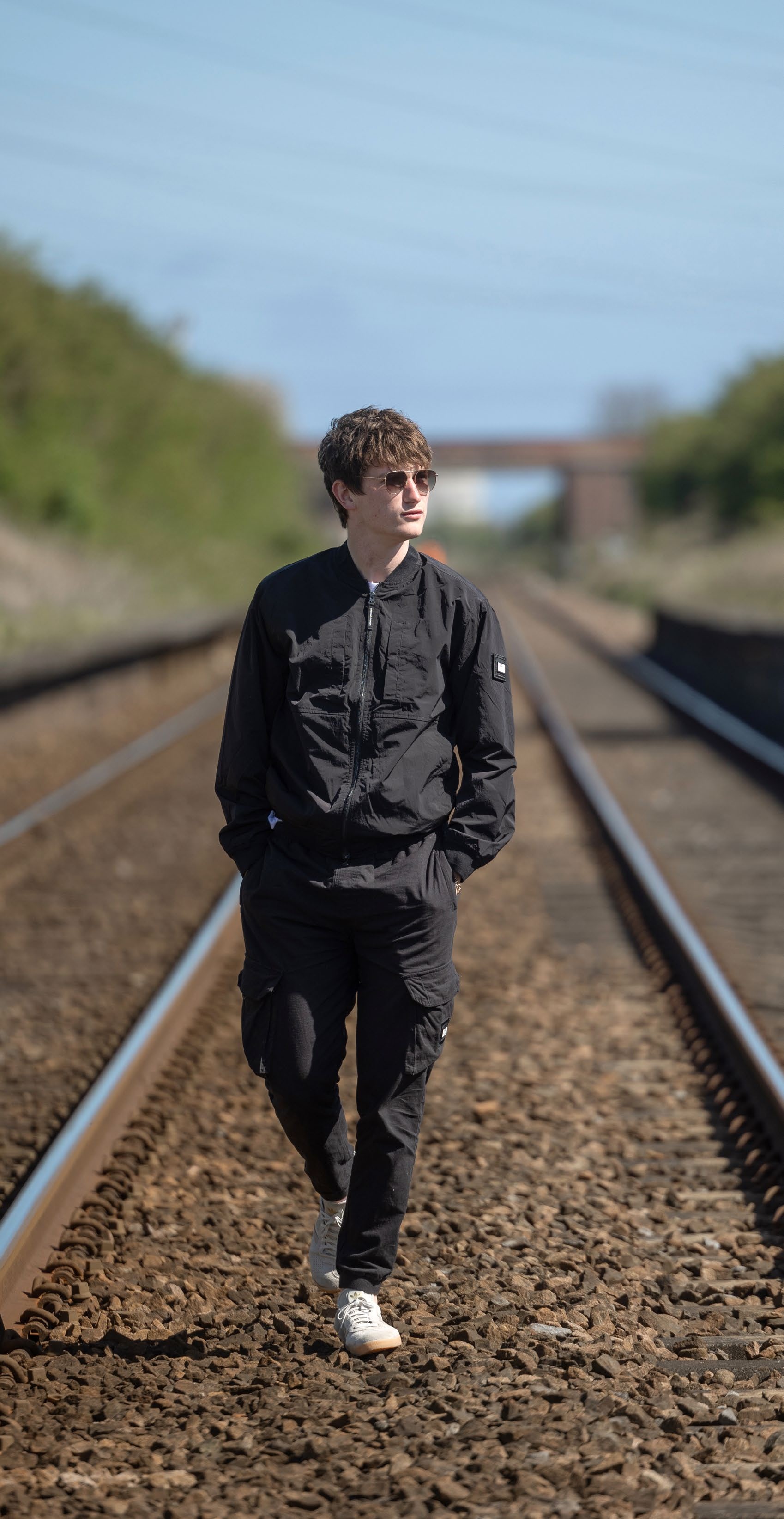 A young man in a black jacket and sunglasses walking along railway tracks, representing a casual and confident style for the FourFourTwo article on Andrew Cushin.