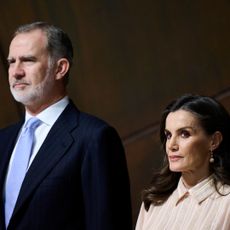 King Felipe of Spain wearing a blue suit and tie standing next to Queen Letizia wearing a peach dress with a collar in front of a brown wall