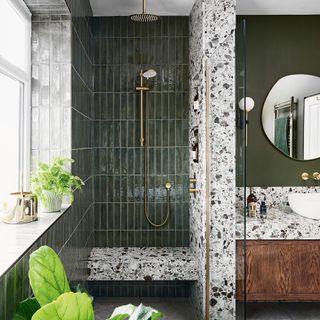 a shower area with dark green tiles and a terrazzo wall and seat beside a florating wooden vanity unit with terrazzo countertop and a white basin