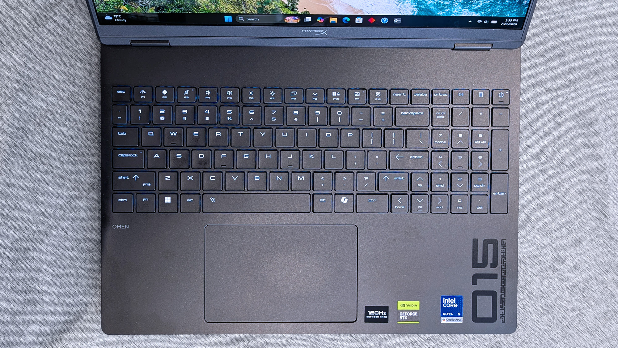 The keyboard and touchpad of a HyperX OMEN 15 laptop, showing a numeric keypad, blue backlit keys, and stickers indicating a 120Hz display, NVIDIA GeForce RTX, and an Intel Core Ultra 9 processor.