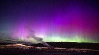 northern lights appear as ribbons of purple and pink light with some green lower down on the horizon across the sky. stream from a geyser rises up in the foreground. 