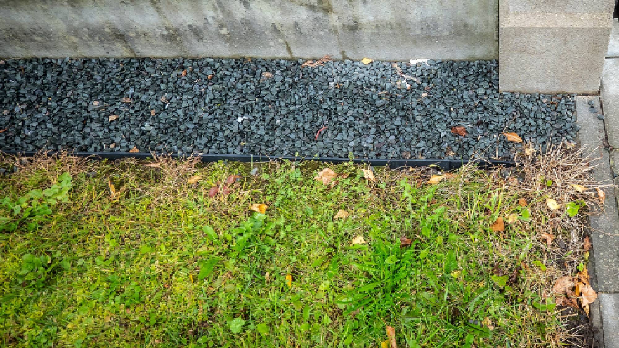 A dark gravel buffer strip running beneath a garden fence to block grass and weeds from spreading.