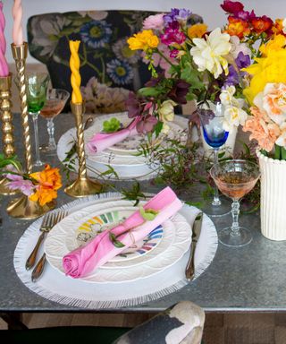 A metal gray dining table with white placemats and plates, pink napkins, gold cutlery and candlesticks with twisted yellow and pink candles, a vase of colorful flowers and colorful glasses, and a floral navy blue chair