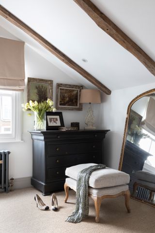 Pippa Jones house: corner of spare bedroom with dark blue-black painted dresser, beige ottoman and large gold-framed mirror, beige carpet and white sloped ceiling with wooden beams