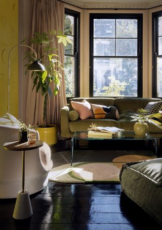 living room with green couch, glass coffee table, black painted floor, plants, bay window, armchairs, side table, floor lamp, black accents