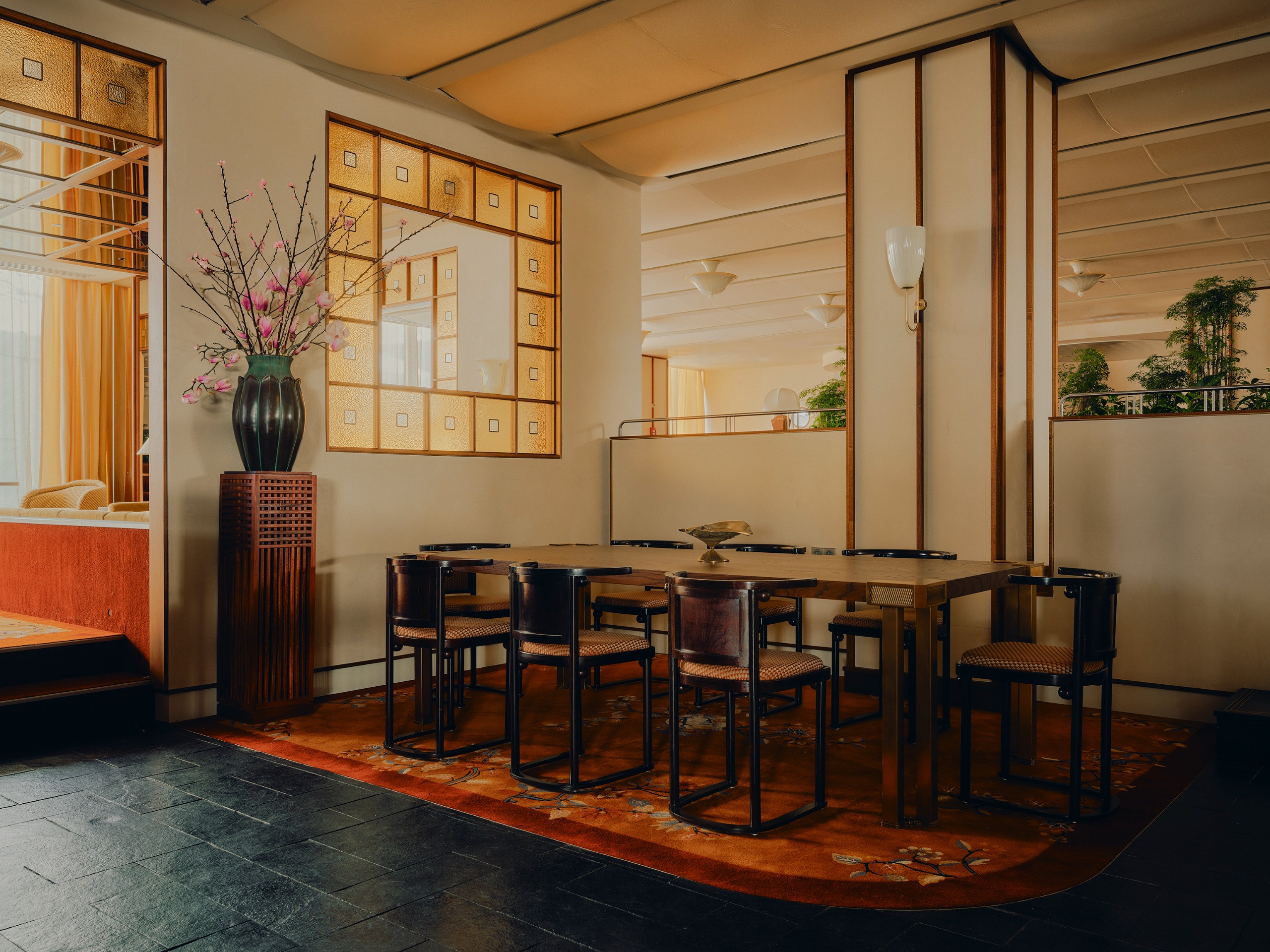 A contemporary restaurant with stained glass internal windows, neutral walls, and a floral carpet underneath a rectangular wooden table with modern chairs