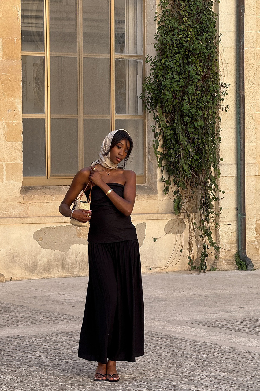 European vacation outfit idea is shown in a photo of a woman standing outside wearing a brown silk headscarf with a black strapless maxi dress, black heeled thong sandals, gold bangles, and a raffia shoulder bag