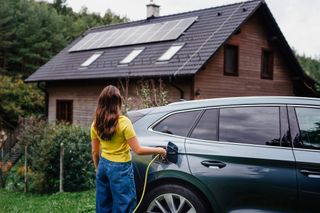 Young woman is charging her electric car, parked in front of a house with solar panels on the roof