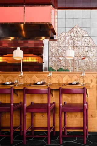 A quirkily decorated ski hotel's restaurant boasts a kitchen counter in wood with bordeaux chairs, brass details, and a tiled mountain-shaped mural.