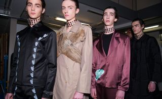 Four male models wearing looks from JW Anderson's collection. The first model is wearing a black jacket and pants with blue detail. The second model is wearing a beige coat with X-shaped fur panel across the chest. The third model is wearing a black crew neck top and light maroon suit with blue detail. And the last model is wearing a black padded coat. All models are wearing thick clear chokers with metal studs