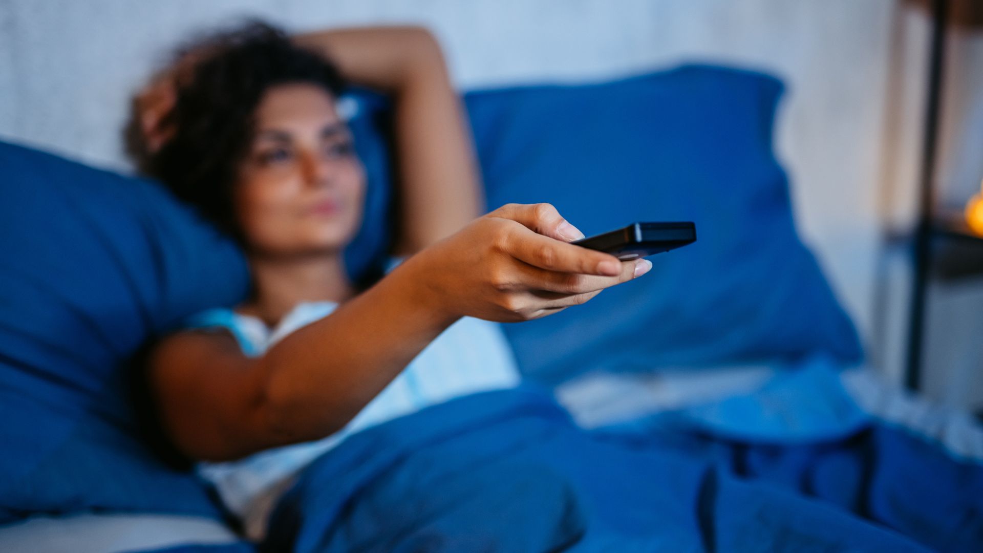 A woman holding a TV remote up watching TV in bed