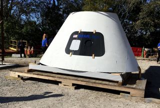 A mock-up of the crew capsule for the reusable CST-100 Starliner, resting under the real Crew Access Arm being built and tested near Kennedy Space Center.