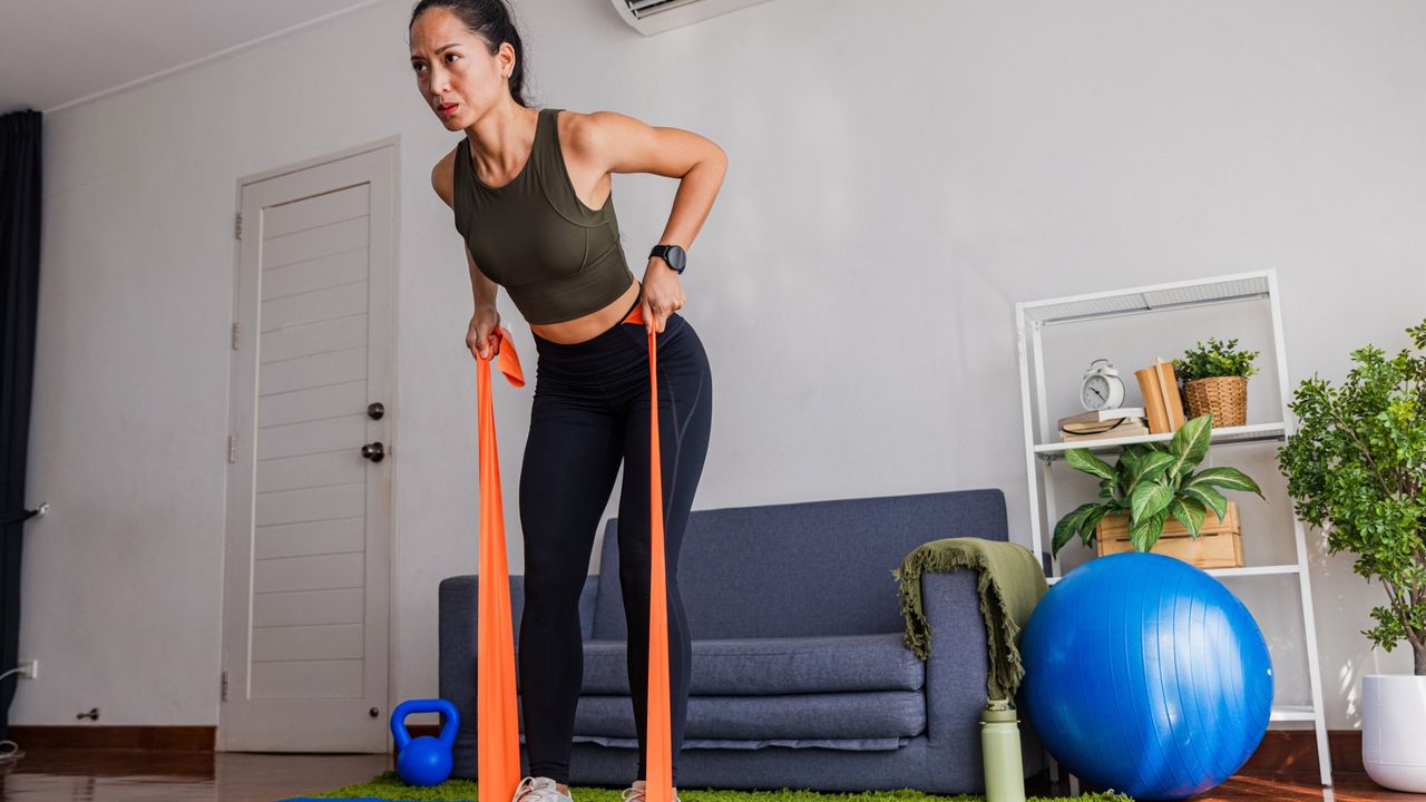 woman in a living room with an orange resistance band under her feet pulling the ends towards her in a bent-over row. there's a blue sofa and big blue balance ball next to her. 
