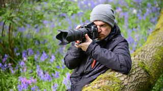 Photographer holding the Nikon D800 DSLR camera up to their eye while leaning on a tree trunk surrounded by bluebells