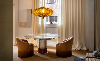 A glass topped round table with two brown chairs, an oval light hanging from above and floor to ceiling curtains. 
