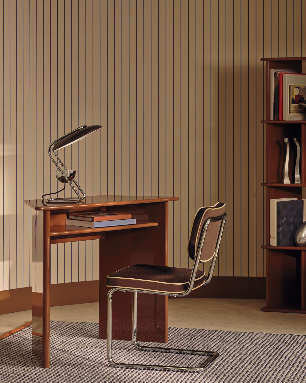 Image of a colorful, mid-century modern style room with a small orange lacquer desk with a brown and metal desk chair. The walls have a cream and navy pinstripe design.