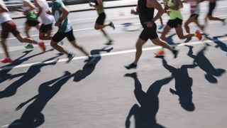 A crowd of people running a road race in the summer, casting long shadows on the street