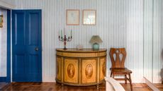 Bedroom with a blue painted door, striped blue wallpaper, a vintage console table with a painted design, an iron candelabra on top with colourful candles, and a vintage chair beside it